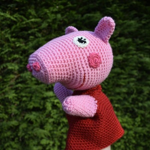 Può includere: Un pupazzo a uncinetto rosa di un maiale che indossa un vestito rosso. Il maiale ha un occhio nero e le guance rosa.