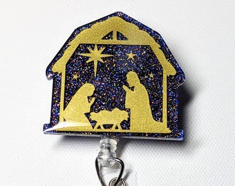 May include: A gold glitter Christmas nativity scene with a blue glitter background. The scene is attached to a clear plastic retractable badge holder.