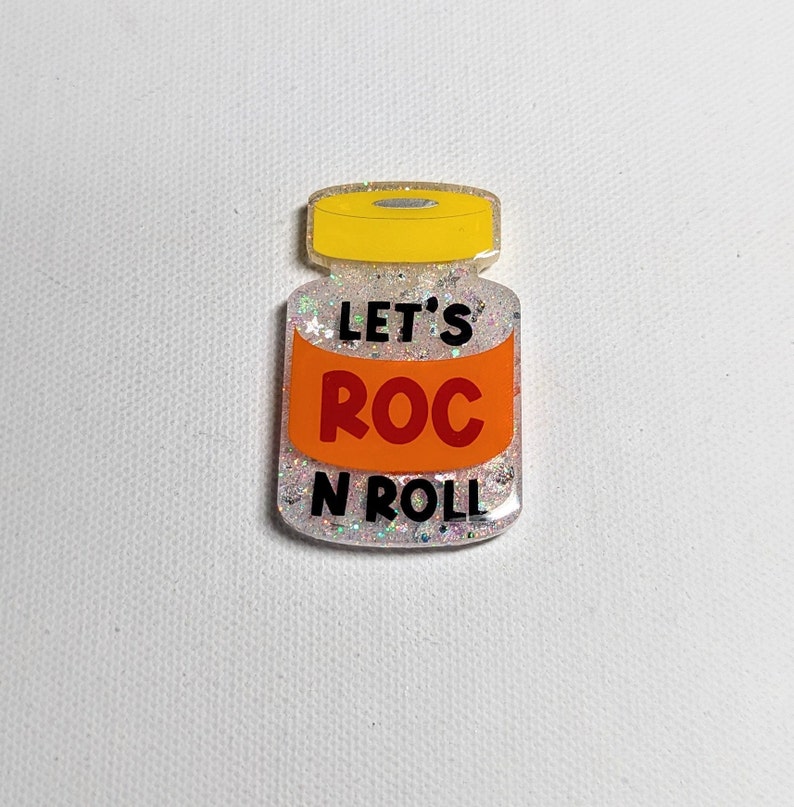 May include: A glittery, jar-shaped pin with a yellow lid and an orange label. The label reads "LET'S ROC N ROLL" in black and white text. The jar is filled with silver glitter.