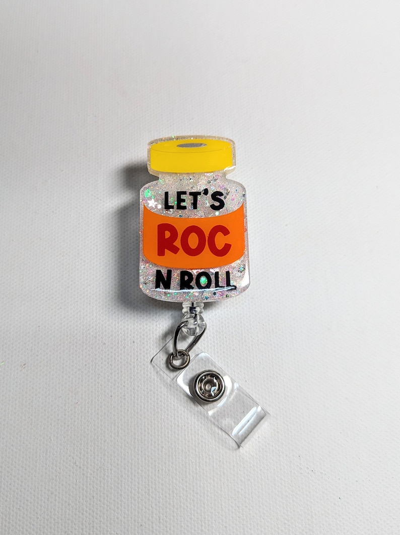 May include: A glittery badge reel in the shape of a vaccine vial. The vial has a yellow cap and an orange label with the words "LET'S ROC N ROLL" in black. The reel has a clear plastic clip.