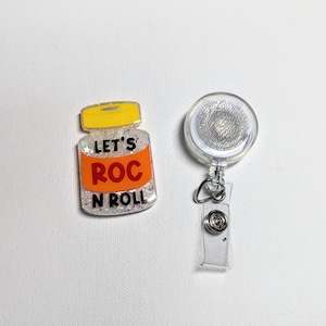May include: A clear badge reel with a silver glitter design and a clear plastic strap. Next to it is a badge with the text "LET'S ROC N ROLL" on an orange background, with a yellow lid and glitter.