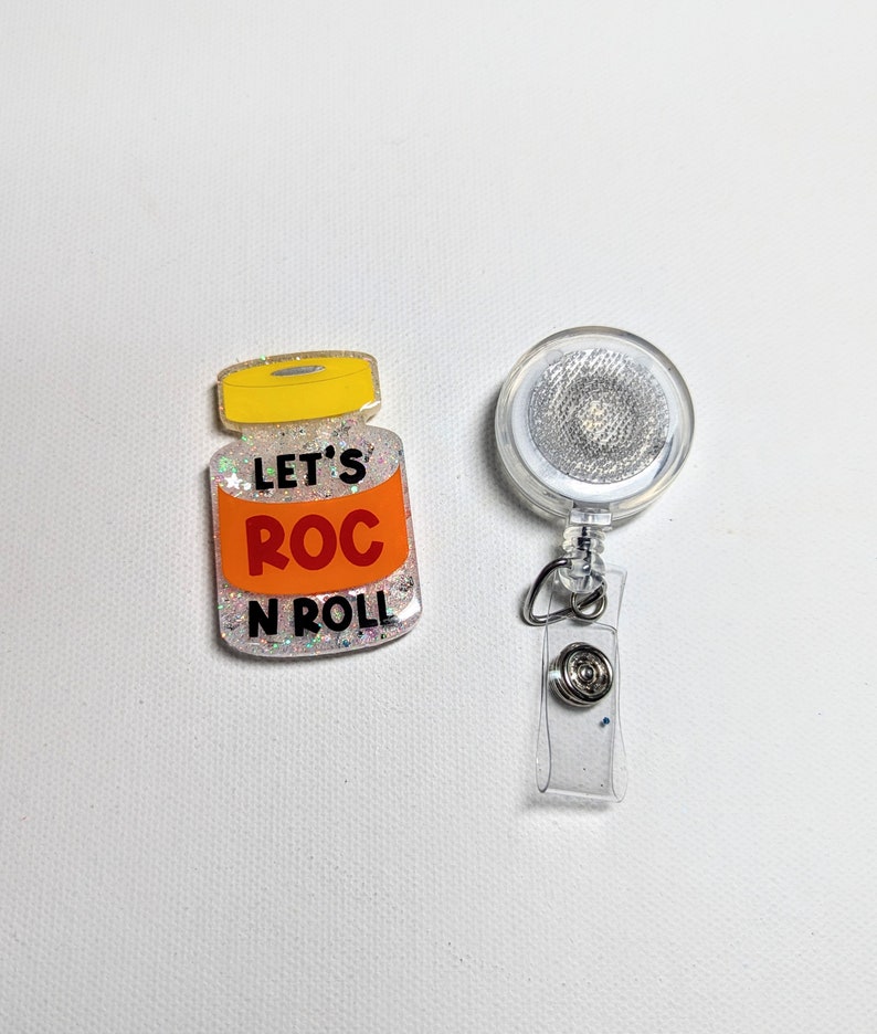 May include: A clear badge reel with a silver glitter design and a clear plastic strap. Next to it is a badge with the text "LET'S ROC N ROLL" on an orange background, with a yellow lid and glitter.