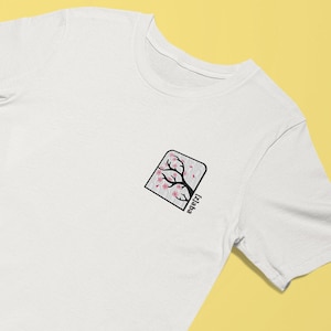 May include: White t-shirt with a small embroidered design of a cherry blossom tree in a square frame. The text "Japan" is written vertically below the tree.