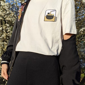 Puede incluir: Camiseta blanca con una imagen bordada en negro y marr&oacute;n de un cuenco de fideos ramen con el texto "Izakaya" en la esquina inferior derecha.