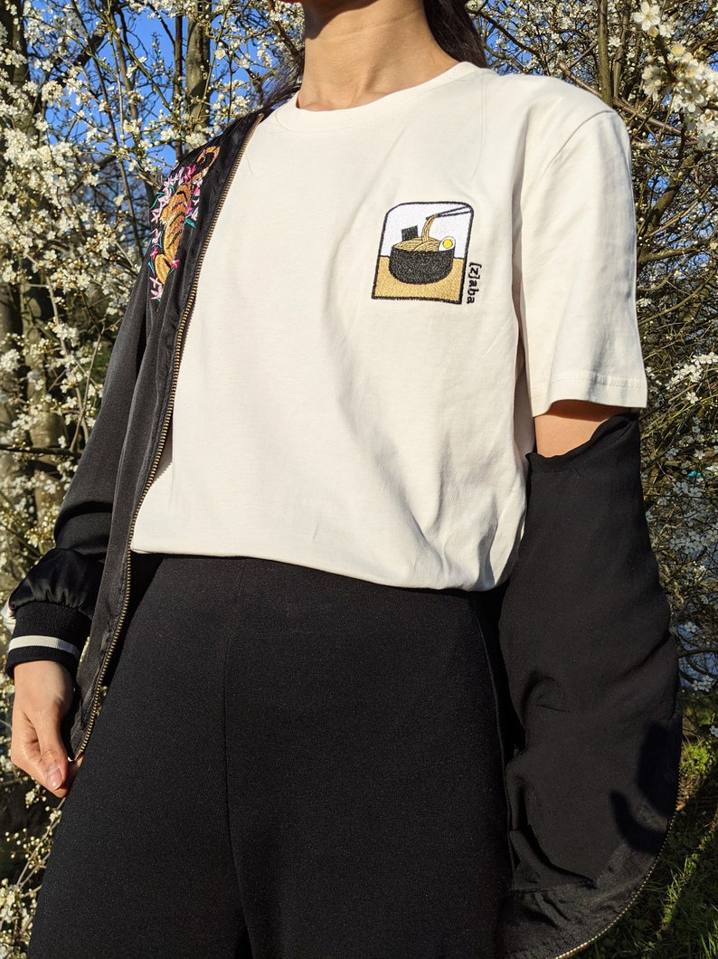 Puede incluir: Camiseta blanca con una imagen bordada en negro y marr&oacute;n de un cuenco de fideos ramen con el texto "Izakaya" en la esquina inferior derecha.