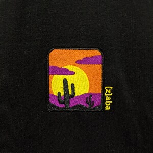 K&ouml;nnte beinhalten: Ein schwarzer gestickter Patch mit einer Sonnenuntergangsszene mit einer gelben Sonne, lila Wolken und zwei lila Kakteen. Der Text "Zaba" ist in Gelb auf der rechten Seite des Patches gestickt.