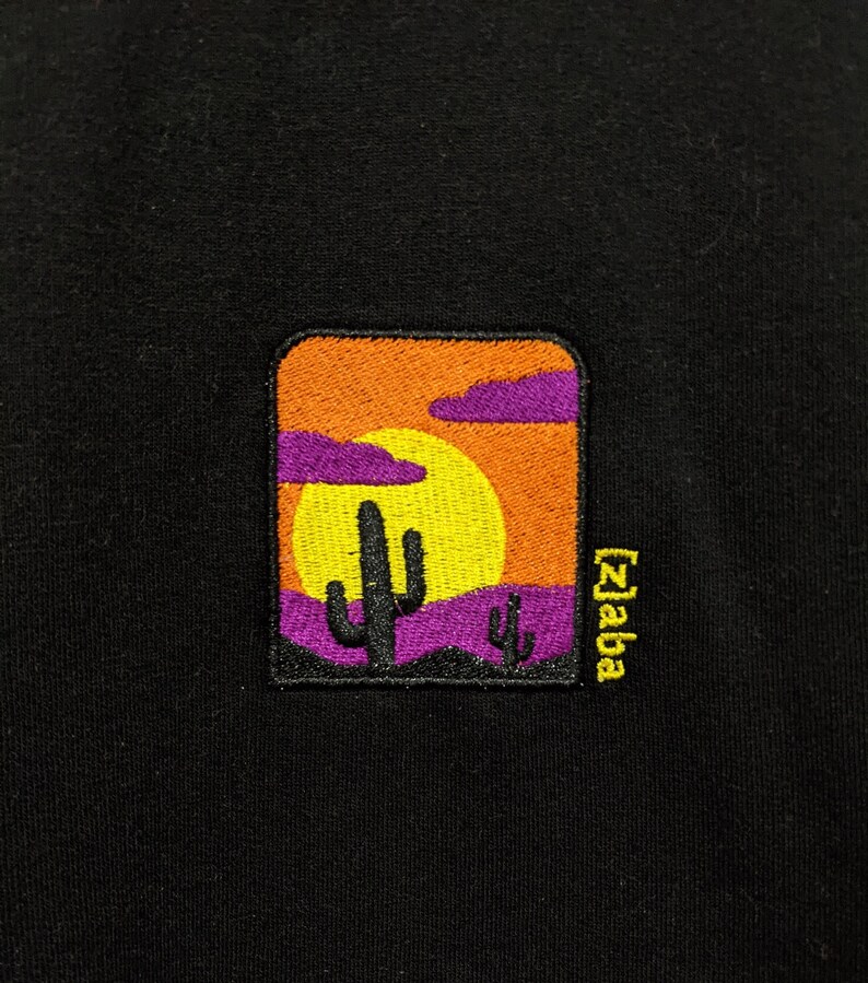 K&ouml;nnte beinhalten: Ein schwarzer gestickter Patch mit einer Sonnenuntergangsszene mit einer gelben Sonne, lila Wolken und zwei lila Kakteen. Der Text "Zaba" ist in Gelb auf der rechten Seite des Patches gestickt.
