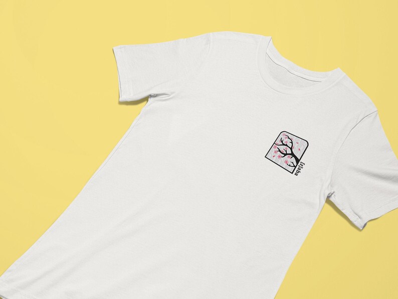 May include: White t-shirt with a small embroidered design of a cherry blossom tree in a square frame. The text "Japan" is written vertically below the tree.