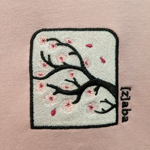 May include: A pink sweatshirt with an embroidered image of a cherry blossom tree branch with pink flowers and the text "[2]aba" in black.