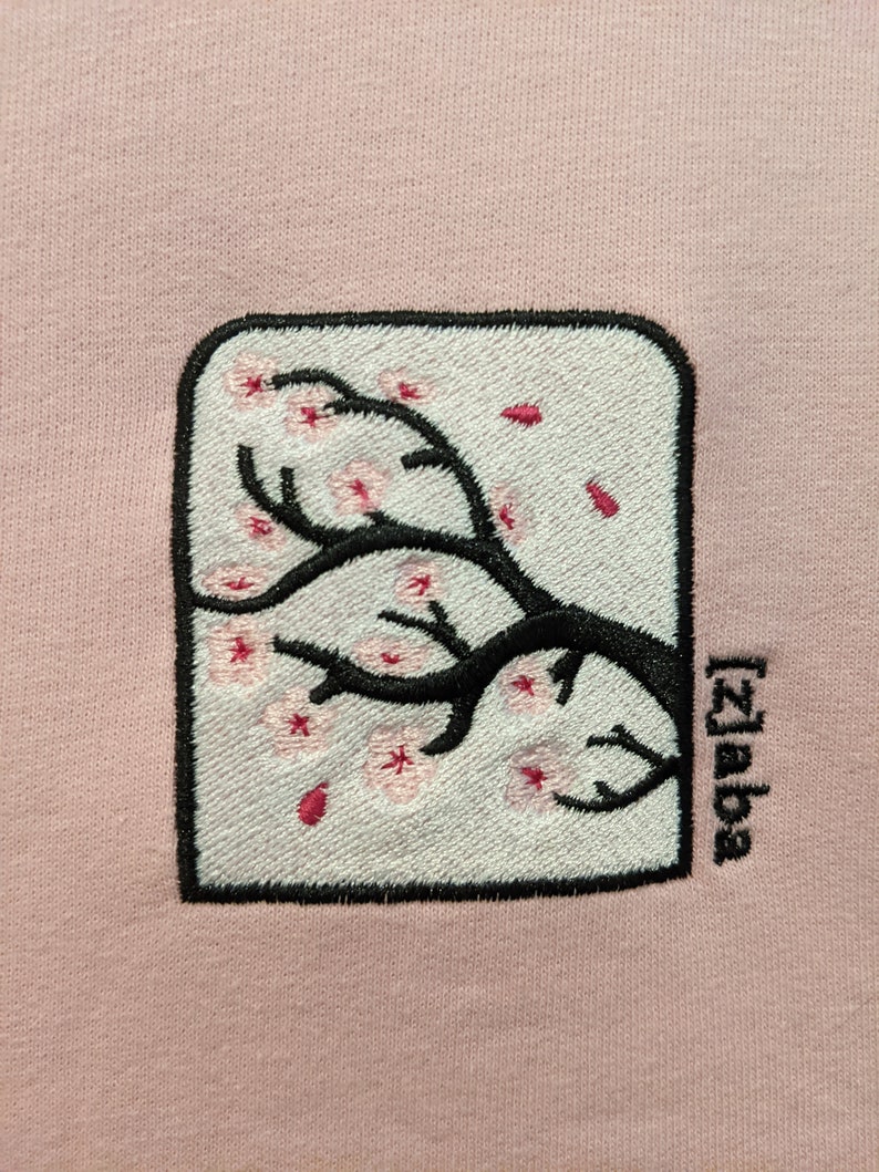 May include: A pink sweatshirt with an embroidered image of a cherry blossom tree branch with pink flowers and the text "[2]aba" in black.