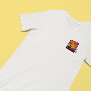 K&ouml;nnte beinhalten: Wei&szlig;es T-Shirt mit einer lila, orangefarbenen und gelben Grafik eines Sonnenuntergangs und eines Kaktus mit dem Text "Arizona" auf der Vorderseite.