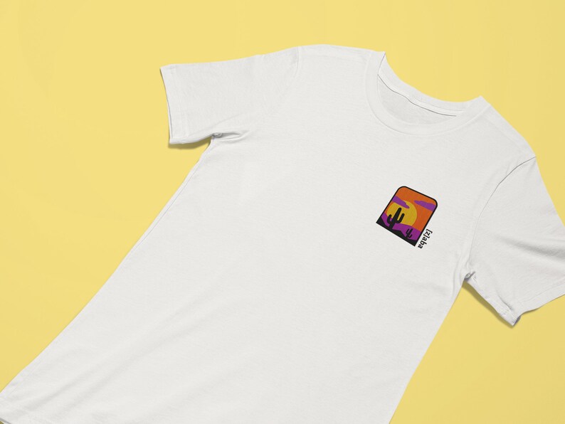 K&ouml;nnte beinhalten: Wei&szlig;es T-Shirt mit einer lila, orangefarbenen und gelben Grafik eines Sonnenuntergangs und eines Kaktus mit dem Text "Arizona" auf der Vorderseite.