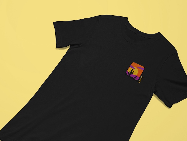 K&ouml;nnte beinhalten: Schwarzes T-Shirt mit einem kleinen gestickten Aufn&auml;her auf der linken Brust. Der Aufn&auml;her zeigt eine Sonnenuntergangsszene mit einem Kaktus und dem Wort "Arizona" in lila Schrift.