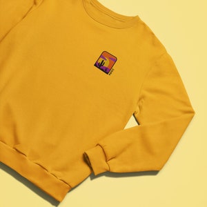 May include: A mustard yellow sweatshirt with a small embroidered patch on the left chest. The patch features a sunset scene with a cactus and the word "Mojave" in purple.