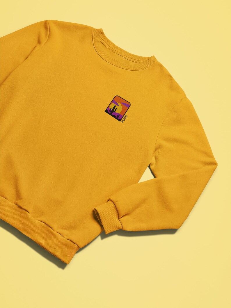 May include: A mustard yellow sweatshirt with a small embroidered patch on the left chest. The patch features a sunset scene with a cactus and the word "Mojave" in purple.