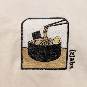 Puede incluir: Dise&ntilde;o bordado de un cuenco de fideos ramen con palillos, un huevo y una alga. El dise&ntilde;o est&aacute; sobre un fondo beige e incluye el texto "[Zaba]" en la esquina inferior derecha.