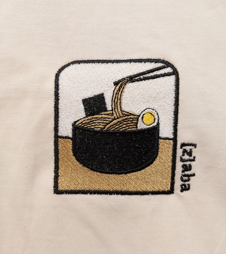 Puede incluir: Dise&ntilde;o bordado de un cuenco de fideos ramen con palillos, un huevo y una alga. El dise&ntilde;o est&aacute; sobre un fondo beige e incluye el texto "[Zaba]" en la esquina inferior derecha.