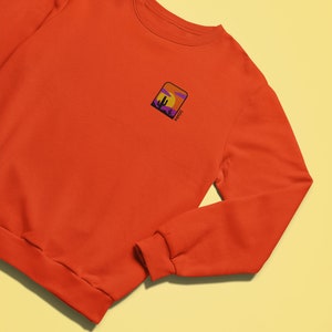 May include: Orange sweatshirt with a small embroidered patch on the left chest. The patch features a sunset scene with a cactus and the word "Sunset" written in a script font.
