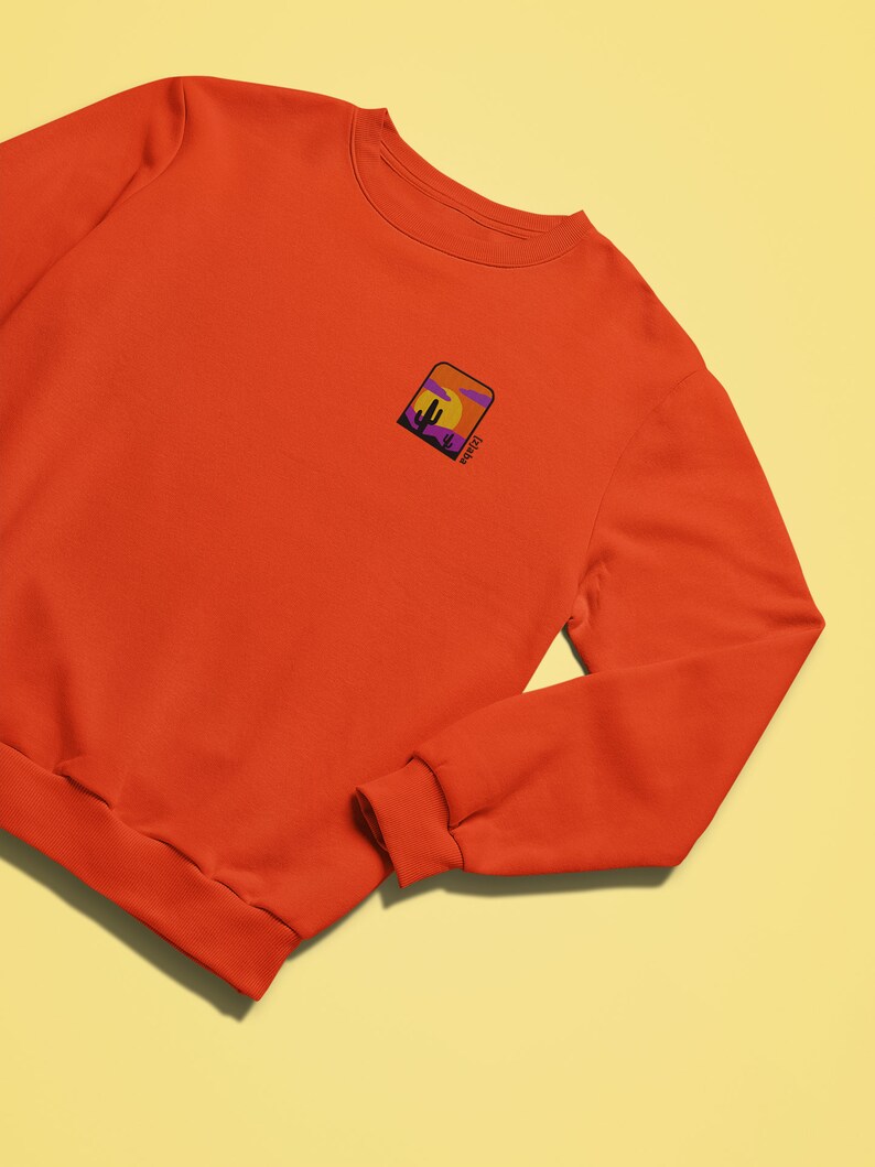 May include: Orange sweatshirt with a small embroidered patch on the left chest. The patch features a sunset scene with a cactus and the word "Sunset" written in a script font.