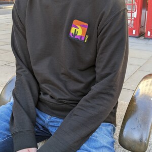 May include: A black long-sleeved sweatshirt with a colorful embroidered patch on the left chest. The patch depicts a desert scene with a cactus, a setting sun, and the text "Cacti".
