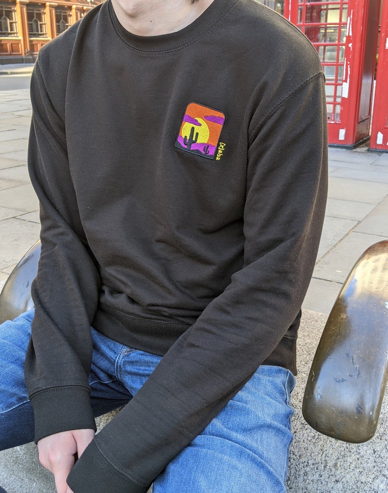May include: A black long-sleeved sweatshirt with a colorful embroidered patch on the left chest. The patch depicts a desert scene with a cactus, a setting sun, and the text "Cacti".