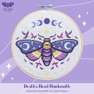 Puede incluir: Patrón de punto de cruz de una esfinge de la calavera, con una calavera, medias lunas y estrellas. Las alas de la polilla son azules con lunas y estrellas blancas, y el cuerpo es morado. El diseño está en un bastidor de bordado de madera.