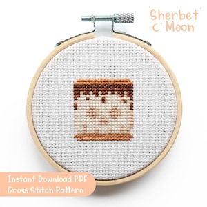 May include: A cross-stitch design of a dessert, presented in a wooden embroidery hoop. The pattern shows a brown top and base with a beige centre. The text "Instant Download PDF Cross Stitch Pattern" is visible.