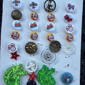 May include: A collection of assorted buttons arranged on a white card with handwritten prices. The set includes round buttons with printed designs of airplanes, trains, clowns, and bicycles, alongside metallic, clear, and green gardening angel shapes.
