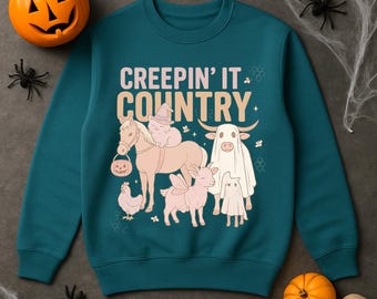 May include: Teal sweatshirt with the phrase "CREEPIN' IT COUNTRY" and illustrations of a horse, cat, cow, goat, and chicken in Halloween costumes. A carved pumpkin, spiders, and a skull are in the background.