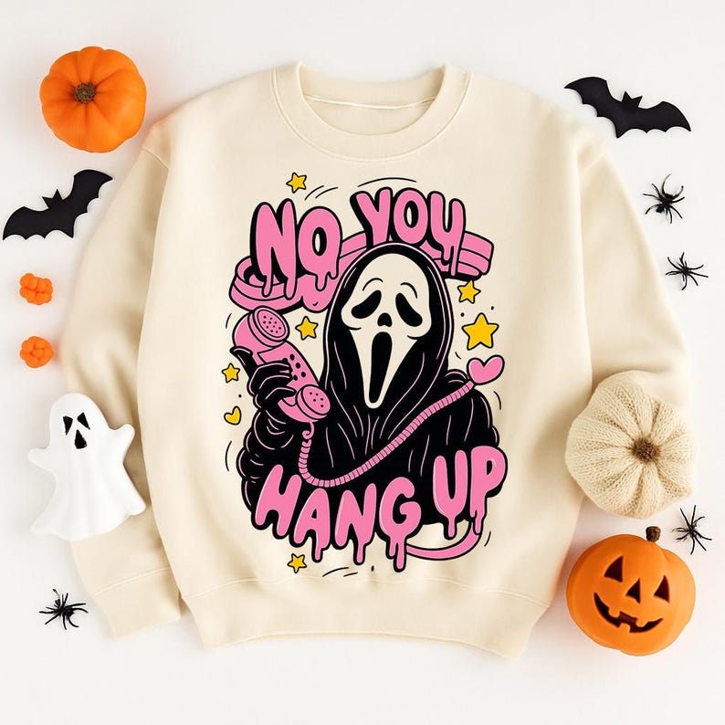 Puede incluir: Sudadera crema con un dise&ntilde;o gr&aacute;fico de la m&aacute;scara de Scream sosteniendo un tel&eacute;fono rosa. El texto "NO YOU HANG UP" est&aacute; en letras rosas. Tem&aacute;tica de Halloween con calabazas, murci&eacute;lagos y ara&ntilde;as.