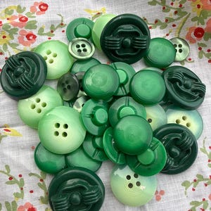 May include: A collection of assorted round buttons in various shades of green, ranging from light mint to deep forest green. The buttons feature different designs, including smooth surfaces, four-hole centres, and raised geometric patterns.