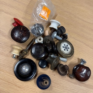 Puede incluir: Una colección de pomos y tiradores de cajón variados en diversos materiales, incluyendo madera oscura, cerámica blanca, plástico negro y metal. Algunos pomos presentan patrones decorativos, texturas circulares o detalles metálicos.