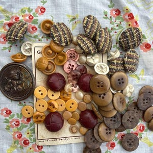 May include: A collection of assorted buttons in various sizes, shapes, and colors, including textured brown fabric-covered buttons, smooth wooden buttons, and a large metallic button featuring an embossed cherub, resting on a vintage Filhar Product card.