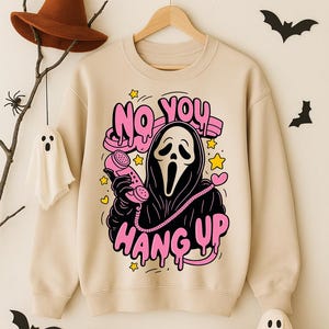 Puede incluir: Sudadera beige con un gr&aacute;fico de un personaje tipo Ghostface sosteniendo un tel&eacute;fono rosa, con las palabras "NO YOU HANG UP" en rosa. Dise&ntilde;o tem&aacute;tico de Halloween con estrellas y corazones. Murci&eacute;lagos decorativos, calabazas y un fantasma en el fondo.