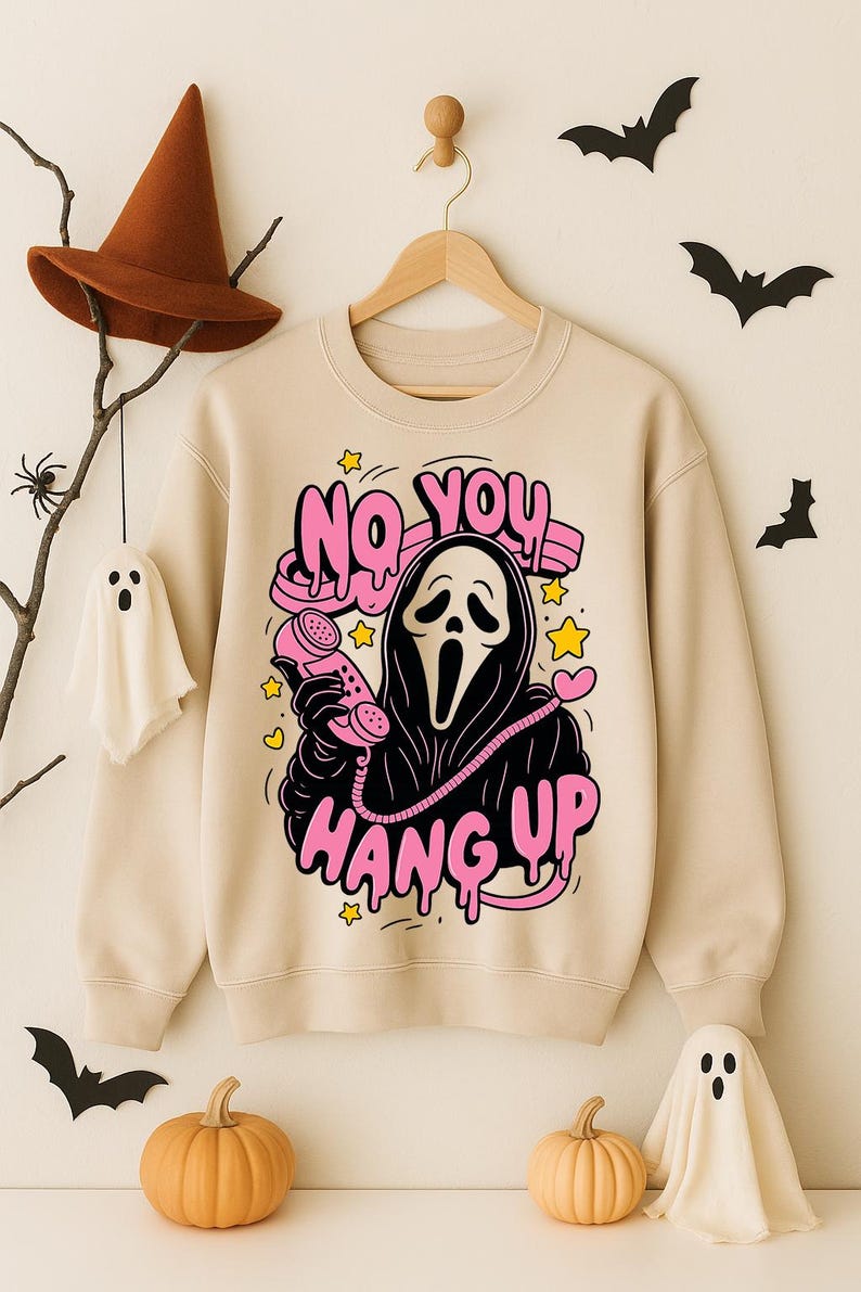 Puede incluir: Sudadera beige con un gr&aacute;fico de un personaje tipo Ghostface sosteniendo un tel&eacute;fono rosa, con las palabras "NO YOU HANG UP" en rosa. Dise&ntilde;o tem&aacute;tico de Halloween con estrellas y corazones. Murci&eacute;lagos decorativos, calabazas y un fantasma en el fondo.
