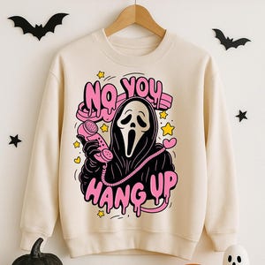 Puede incluir: Sudadera color crema con un gr&aacute;fico de la m&aacute;scara de Ghostface de las pel&iacute;culas Scream sosteniendo un tel&eacute;fono rosa. El texto "NO YOU HANG UP" est&aacute; encima del personaje, y "HANG UP" est&aacute; debajo. Decoraciones tem&aacute;ticas de Halloween rodean la sudadera.