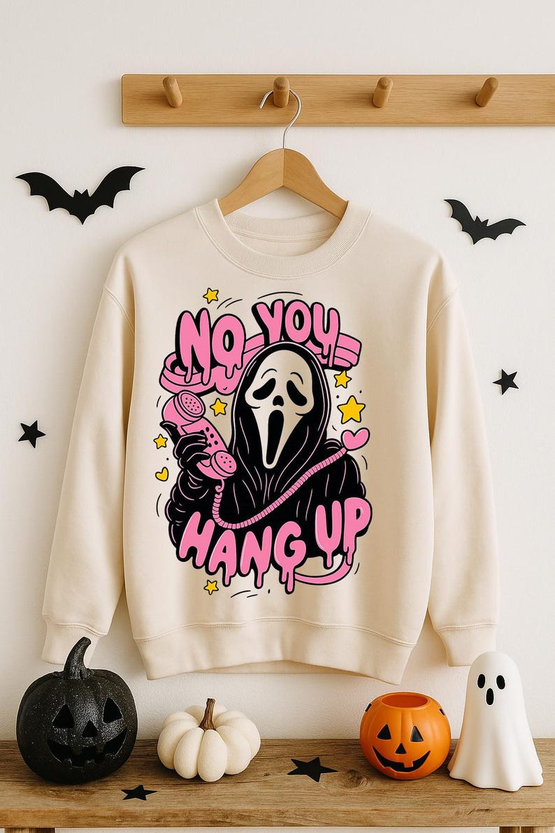 Puede incluir: Sudadera color crema con un gr&aacute;fico de la m&aacute;scara de Ghostface de las pel&iacute;culas Scream sosteniendo un tel&eacute;fono rosa. El texto "NO YOU HANG UP" est&aacute; encima del personaje, y "HANG UP" est&aacute; debajo. Decoraciones tem&aacute;ticas de Halloween rodean la sudadera.