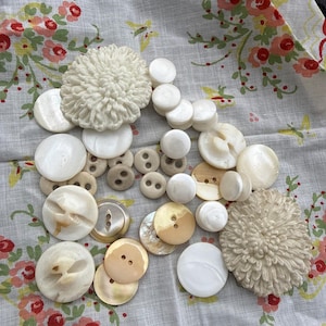 May include: A collection of assorted white and cream buttons in various sizes and shapes, including two large textured floral-shaped buttons, several smooth round buttons, and some with two or four holes, scattered across a white fabric surface.