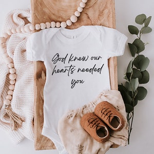 May include: White baby onesie with the text "God knew our hearts needed you" in black script font. The onesie is laid out on a wooden tray with a white blanket, wooden beads, and a sprig of eucalyptus.