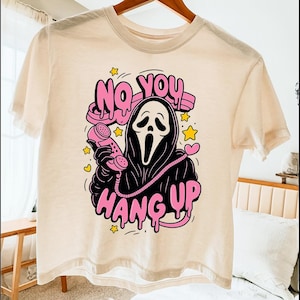 Puede incluir: Camiseta color crema con un gr&aacute;fico del asesino Ghostface de las pel&iacute;culas Scream. El dise&ntilde;o incluye las frases "NO YOU" y "HANG UP" en rosa, con estrellas y corazones amarillos. El personaje sostiene un tel&eacute;fono rosa.