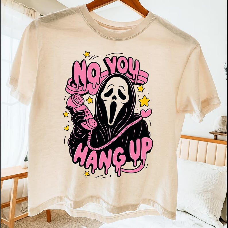 Puede incluir: Camiseta color crema con un gr&aacute;fico del asesino Ghostface de las pel&iacute;culas Scream. El dise&ntilde;o incluye las frases "NO YOU" y "HANG UP" en rosa, con estrellas y corazones amarillos. El personaje sostiene un tel&eacute;fono rosa.