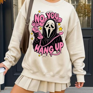Puede incluir: Sudadera de color crema con un gr&aacute;fico de la m&aacute;scara de Ghostface de las pel&iacute;culas Scream sosteniendo un tel&eacute;fono rosa. El texto "NO YOU HANG UP" est&aacute; encima de la m&aacute;scara y "HANG UP" debajo, en letras rosas.