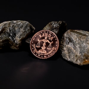 May include: A copper coin displaying the Libra zodiac symbol, a balanced scale, with the word "LIBRA" above. The coin also features the dates "September 24 - October 23". The coin is set against a dark background with rough, grey stones.