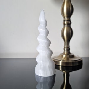 Puede incluir: Una torre de cristal de selenita blanca con un diseño en espiral. El cristal se coloca sobre una superficie negra reflectante, con una lámpara de latón en el fondo. La torre mide aproximadamente 15 cm de altura.