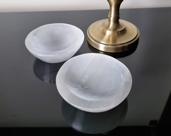May include: Three white selenite bowls with a smooth, translucent appearance are arranged on a reflective black surface. A brass-colored candlestick holder is partially visible in the background, adding a touch of contrast.