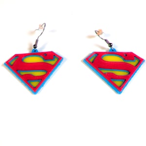 May include: A pair of Superman logo earrings. Each earring features the iconic Superman logo in red, yellow, and blue. The earrings have a blue outline, a red diamond shape, and a yellow center. The earrings hang from silver-colored fish hook ear wires.