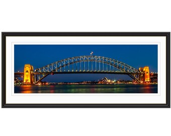 May include: A panoramic view of the Sydney Harbour Bridge at night, illuminated with warm yellow lights against a dark blue sky. The bridge is a steel arch bridge with a distinctive arch shape.