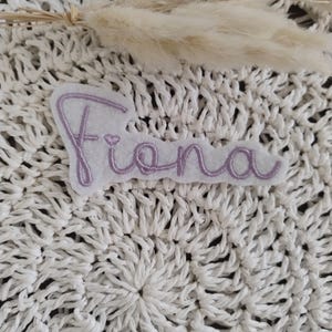 Könnte beinhalten: Ein weißes Filz-Patch mit dem Namen "Fiona" in einer kursiven, hellvioletten Schriftart gestickt. Der Patch befindet sich auf einer strukturierten, weißen, gewebten Oberfläche, mit getrocknetem Ziergras im Hintergrund.