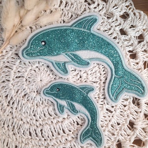 Könnte beinhalten: Zwei türkisfarbene Glitzer-Delfin-Patches, einer größer als der andere, auf einer weißen Häkel-Oberfläche.