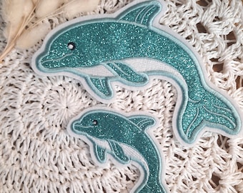 Könnte beinhalten: Zwei türkisfarbene Glitzer-Delfin-Patches, einer größer als der andere, auf einer weißen Häkel-Oberfläche.